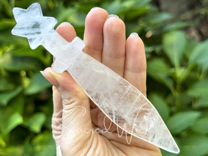 Clear quartz crystal athame with star handle held between fingers, greenery in the background.