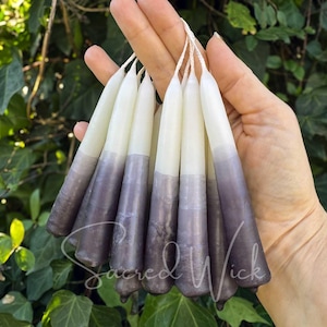 May include: A bunch of taper candles with a white top and a gradient to a dark purple color. The candles are held in a hand with the text "Sacred Wick" visible at the bottom of the image.