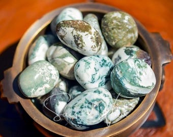 May include: A collection of polished, smooth stones in a bronze-colored bowl. The stones are primarily white with green and brown patterns. The bowl has a rustic, aged appearance, and the background is a warm, orange-brown.