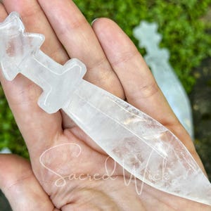 Another clear quartz crystal athame resting on open palm, sunlight with greenery and stones behind.