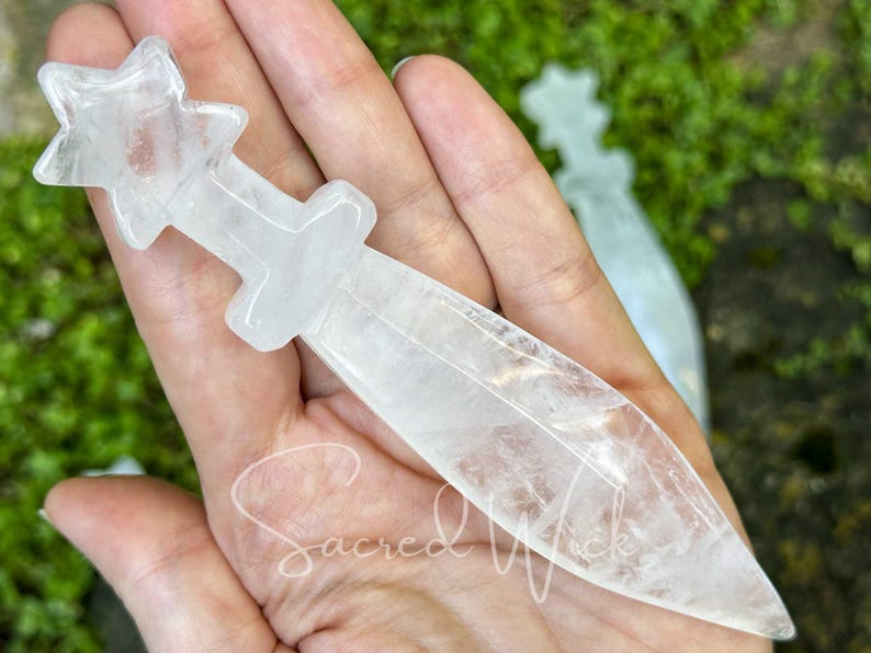 Another clear quartz crystal athame resting on open palm, sunlight with greenery and stones behind.