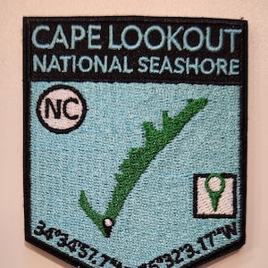 May include: Embroidered patch featuring "CAPE LOOKOUT NATIONAL SEASHORE" in black text on a light blue background. Includes a green island outline, the letters "NC" in a circle, and coordinates. The patch has a black border.
