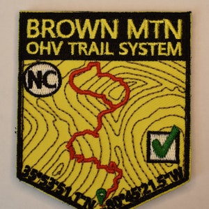 May include: A yellow embroidered patch with black outlining. The patch features the text "BROWN MTN OHV TRAIL SYSTEM" and a map of a trail with a green checkmark. The patch also includes the letters "NC" in a circle.