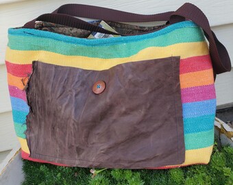 May include: A colorful shoulder bag with a rainbow striped pattern. The bag features a large brown pocket with a wooden button closure. The strap and top trim are brown. The bag is sitting on a bed of green plants.