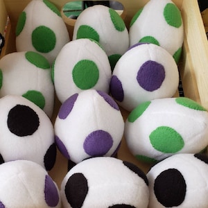 May include: A collection of plush egg-shaped toys in a light-coloured wooden crate. Each white fabric egg features circular felt spots in shades of green, purple, or black, stitched onto the surface. The soft toys are arranged in a group.