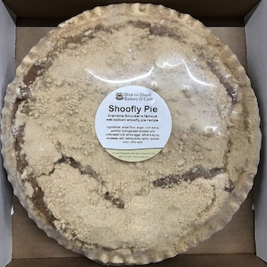May include: A whole Shoofly Pie in a cardboard box, wrapped in clear plastic. The pie has a crumbly, light brown topping and a darker brown crust. A label on the pie reads "Shoofly Pie" and lists ingredients. From Bird-in-Hand Bakery & Cafe.