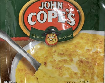 May include: A package of John Cope's Dried Sweet Corn. The green and orange package features the brand's logo and a cartoon image. The image shows a serving of the corn being scooped with a spoon. The net weight is 3.75oz (106g).