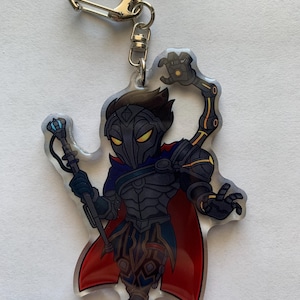 Op de afbeelding: Een sleutelhanger van helder acryl met een zwart en zilveren personage met een rode cape en een mechanische arm die een staf vasthoudt.