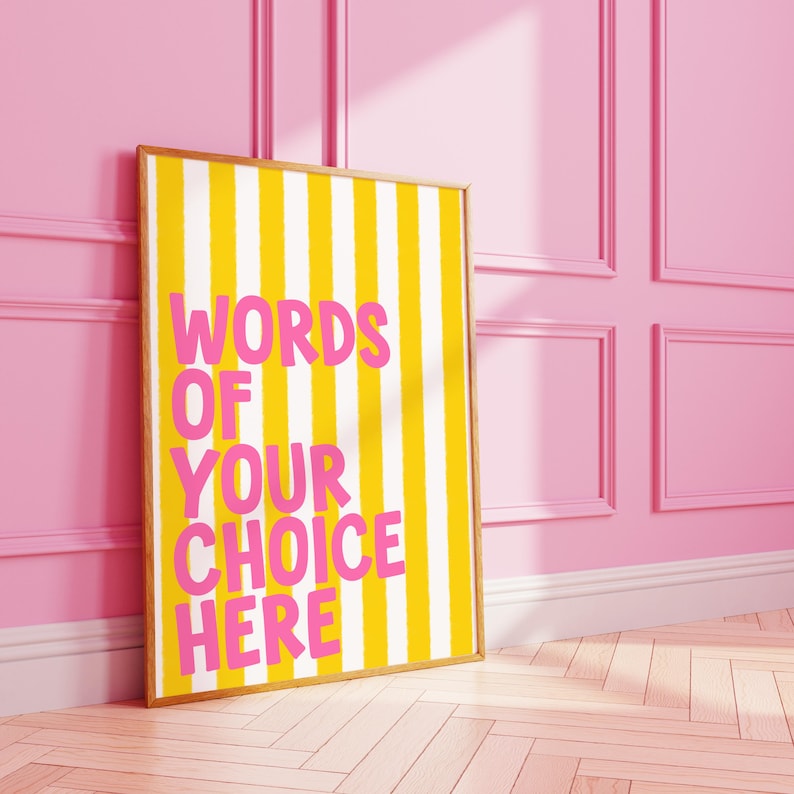 May include: A framed poster with a yellow and white striped background and the text "WORDS OF YOUR CHOICE HERE" in pink. The frame is a light brown color. The poster is leaning against a pink wall.