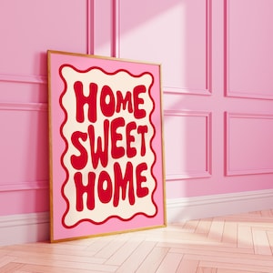 Puede incluir: Un póster rosa y blanco enmarcado con el texto "Home Sweet Home" en letras rojas. El póster está enmarcado en un marco dorado y está apoyado contra una pared rosa.