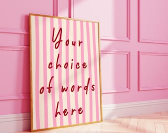 May include: A framed print with a pink and cream striped background. The text "Your choice of words here" is written in a cursive font in red. The frame is gold-colored, and the print is leaning against a pink wall.