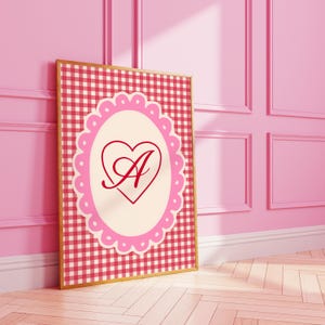 Puede incluir: Un cuadro enmarcado con un diseño de cuadros rosas y rojos. El cuadro tiene un óvalo festoneado rosa claro con un corazón rojo que contiene la letra "A". El marco es dorado y el cuadro está apoyado contra una pared rosa.