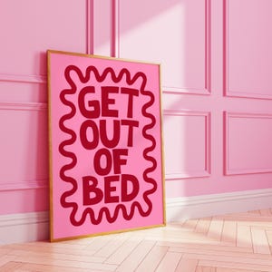 以下が含まれることがあります： 「GET OUT OF BED」の赤い文字が波状の縁で囲まれた、ピンク色の額入りアートプリント。 フレームは金色です。 プリントは、白いトリムと木製の床があるピンク色の壁に立てかけられています。