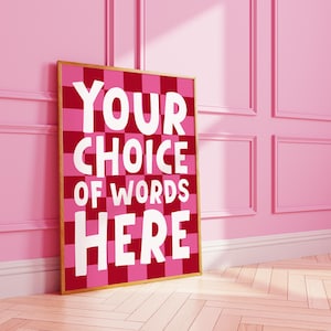 Puede incluir: Un estampado enmarcado con un patrón de tablero de ajedrez rosa y rojo. El estampado presenta las palabras "YOUR CHOICE OF WORDS HERE" en letras blancas grandes. El marco es de color marrón claro y el fondo es una pared rosa.
