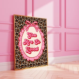 May include: A framed art print with a leopard print background. The print features a pink oval with the words "You Are So Loved" in a retro font. A pink bow and ruffled trim surround the oval. The frame is a light brown colour.