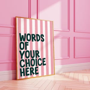 May include: A framed poster with a pink and white striped background. The poster has the words "WORDS OF YOUR CHOICE HERE" in dark green lettering. The frame is a light brown color. The poster is leaning against a pink wall.