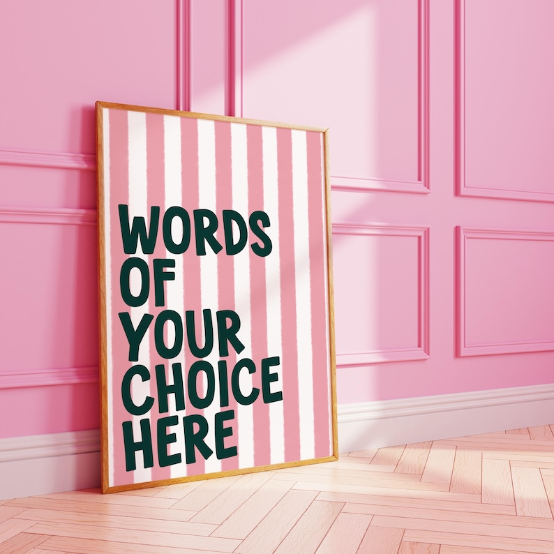 May include: A framed poster with a pink and white striped background. The poster has the words "WORDS OF YOUR CHOICE HERE" in dark green lettering. The frame is a light brown color. The poster is leaning against a pink wall.