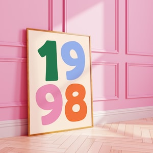 May include: A framed art print featuring the numbers "1998" in bold, colorful typography. The numbers are in green, blue, pink, and orange against a cream background. The artwork is leaning against a pink wall.