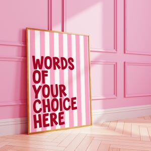 Könnte beinhalten: Ein gerahmter Druck mit einem rosa-weiß gestreiften Hintergrund. Der Text "WORDS OF YOUR CHOICE HERE" ist in roter Schrift dargestellt. Der Rahmen ist goldfarben, und der Druck lehnt an einer rosa Wand.