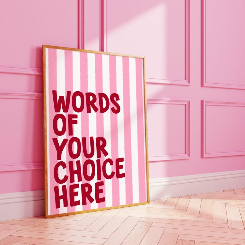 May include: A framed print with a pink and white striped background. The print features the text "WORDS OF YOUR CHOICE HERE" in a bold, red font. The frame is a light gold color, and the print is leaning against a pink wall.