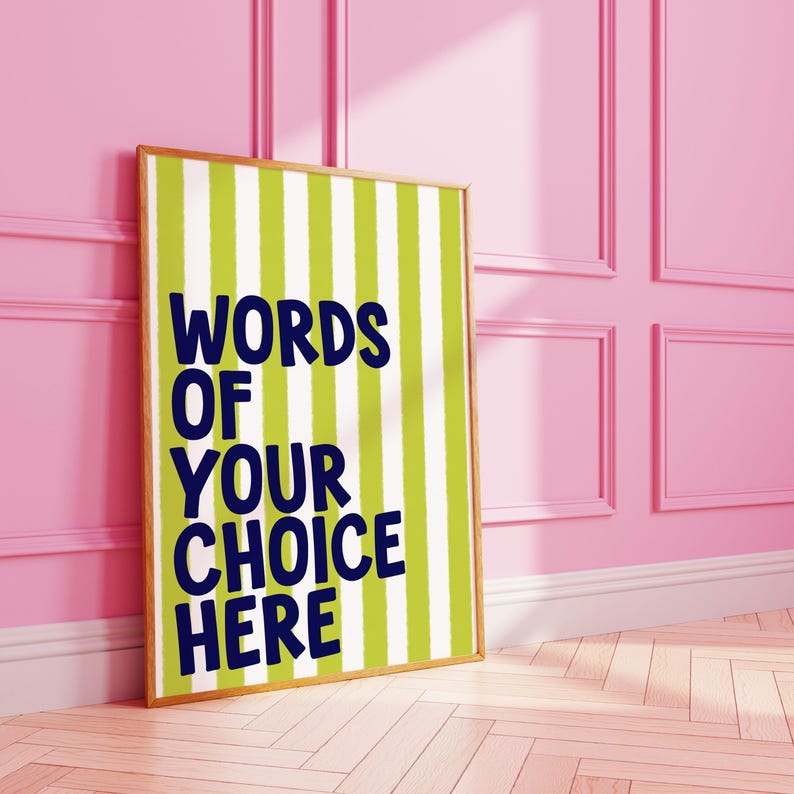 May include: A framed poster with a green and white striped background and the text "WORDS OF YOUR CHOICE HERE" in navy blue. The frame is a light brown color. The poster is leaning against a pink wall.