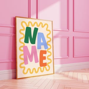 May include: A framed art print with the word "NAME" in colorful, bold letters. The letters are green, blue, pink, and orange, set against a cream background with a yellow wavy border. The artwork is leaning against a pink wall.