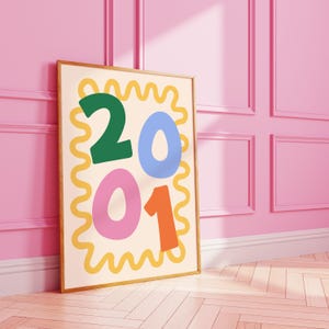 May include: A framed art print featuring the numbers "2001" in bold, colorful fonts. The numbers are in green, blue, pink, and orange, set against a cream background with a yellow wavy border. The artwork is displayed against a pink wall.
