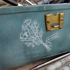 May include: A teal green metal box with a brass latch. The box is open and contains paintbrushes and paint tubes. A white sticker with a floral design and the text "the color joy" is on the inside of the box.