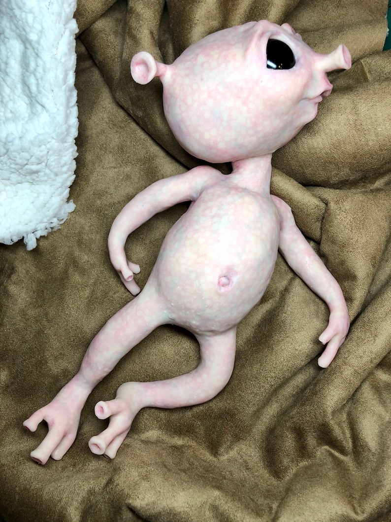 May include: A pale pink alien-like sculpture with large black eyes, small ears, and a protruding nose. The figure has a rounded belly, long arms and legs, and is posed on a brown surface. The sculpture has a textured surface.