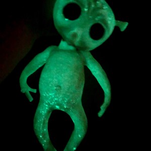 May include: A glowing green alien figurine with large black eyes, small ears, and slender limbs. The toy has a slightly bent posture and is set against a black background, creating a striking contrast. The alien's skin has a speckled texture.