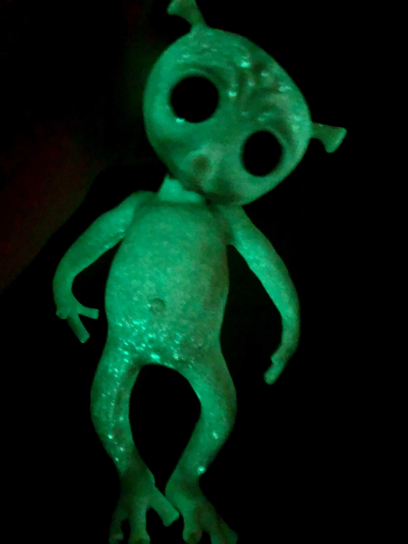 May include: A glowing green alien figurine with large black eyes, small ears, and slender limbs. The toy has a slightly bent posture and is set against a black background, creating a striking contrast. The alien's skin has a speckled texture.