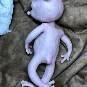 May include: A pale pink, humanoid sculpture with a large head, small arms, and crossed legs. The figure has prominent ears and a smooth, textured surface. It is lying on a brown, textured fabric.
