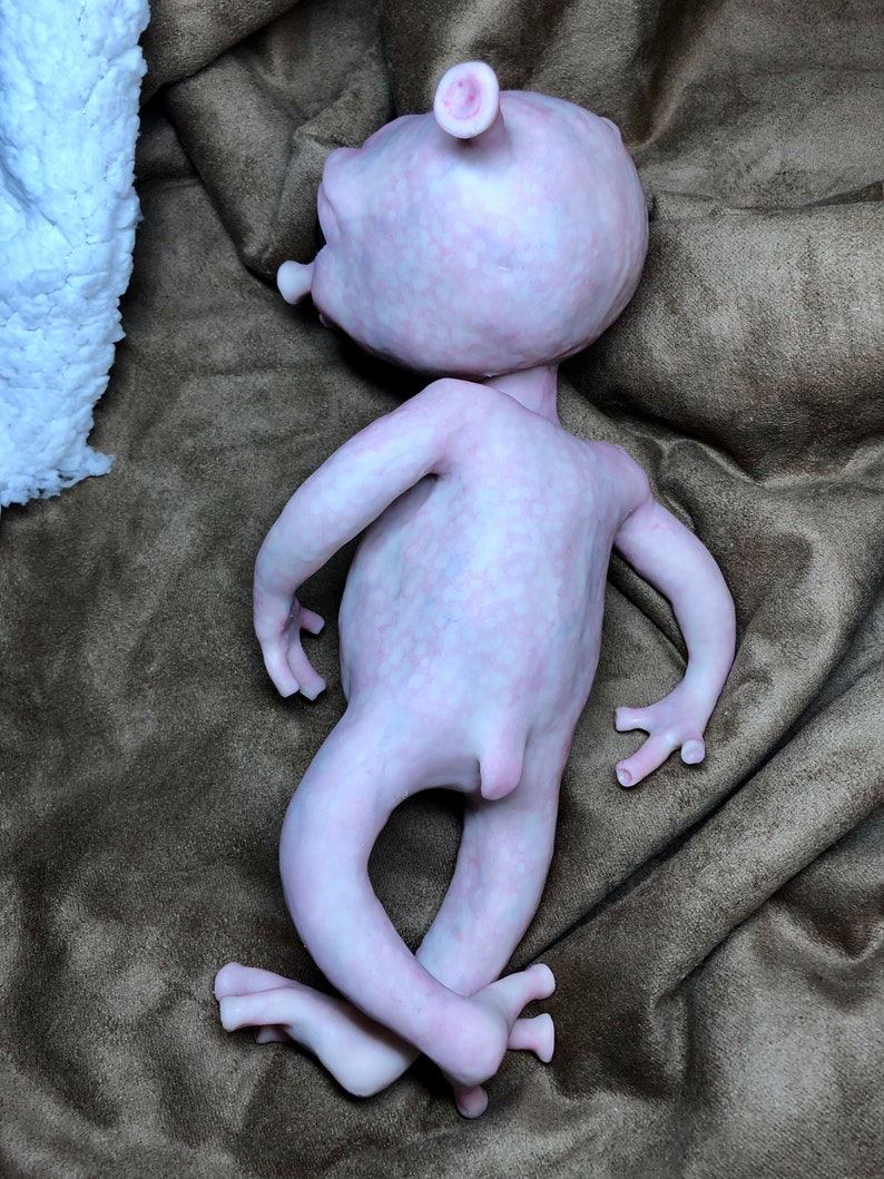May include: A pale pink, humanoid sculpture with a large head, small arms, and crossed legs. The figure has prominent ears and a smooth, textured surface. It is lying on a brown, textured fabric.