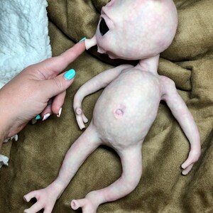 May include: A pale pink alien doll with large black eyes, a small nose, and a single ear. The doll has a smooth, textured skin and long, slender limbs. The doll is lying on a brown surface.