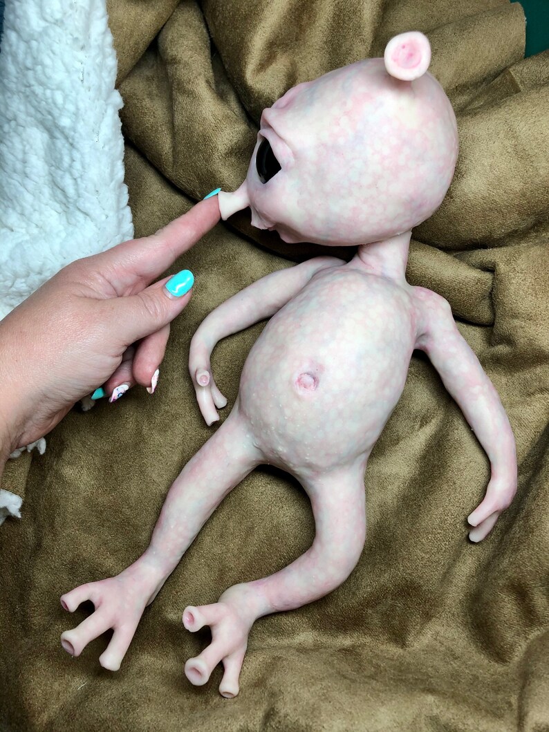 May include: A pale pink alien doll with large black eyes, a small nose, and a single ear. The doll has a smooth, textured skin and long, slender limbs. The doll is lying on a brown surface.