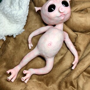 May include: A pale pink, humanoid sculpture with large black eyes, small ears, and a prominent belly button. The figure is lying on a brown fabric surface, with a white, textured material in the background. The sculpture has slender limbs and detailed hands and feet.
