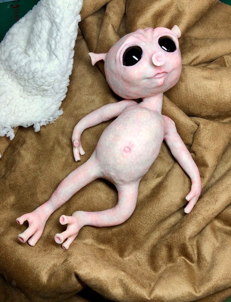 May include: A pale pink, humanoid sculpture with large black eyes, small ears, and a prominent belly button. The figure is lying on a brown fabric surface, with a white, textured material in the background. The sculpture has slender limbs and detailed hands and feet.