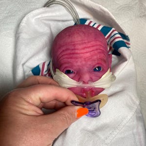 May include: A reborn doll with a pinkish-red complexion, blue eyes, and a pacifier. The doll is wrapped in a white blanket with blue and red stripes. A hand with orange nail polish holds the pacifier.
