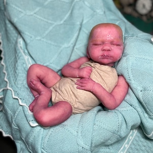 Może przedstawiać: Realistyczna lalka reborn o realistycznym odcieniu skóry, ubrana w beżowe body. Lalka leży na jasnoniebieskim, dzierganym kocu z białym, falistym brzegiem. Lalka ma zamknięte oczy i szczegółowe rysy twarzy.