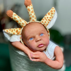 Może przedstawiać: Lalka dla dziecka z jasnoniebieskimi oczami w nakryciu głowy z motywem żyrafy. Nakrycie głowy jest żółte z brązowymi plamami i ma trzy pionowe, cylindryczne kształty. Lalka ma na sobie białe body i jest otulona w miękką, jasną otulinę.