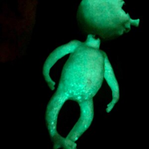 May include: A glowing green alien figurine with a large head and small body. The alien has a small antenna and is posed with its arms outstretched and legs crossed. The figurine has white speckles and is set against a black background.