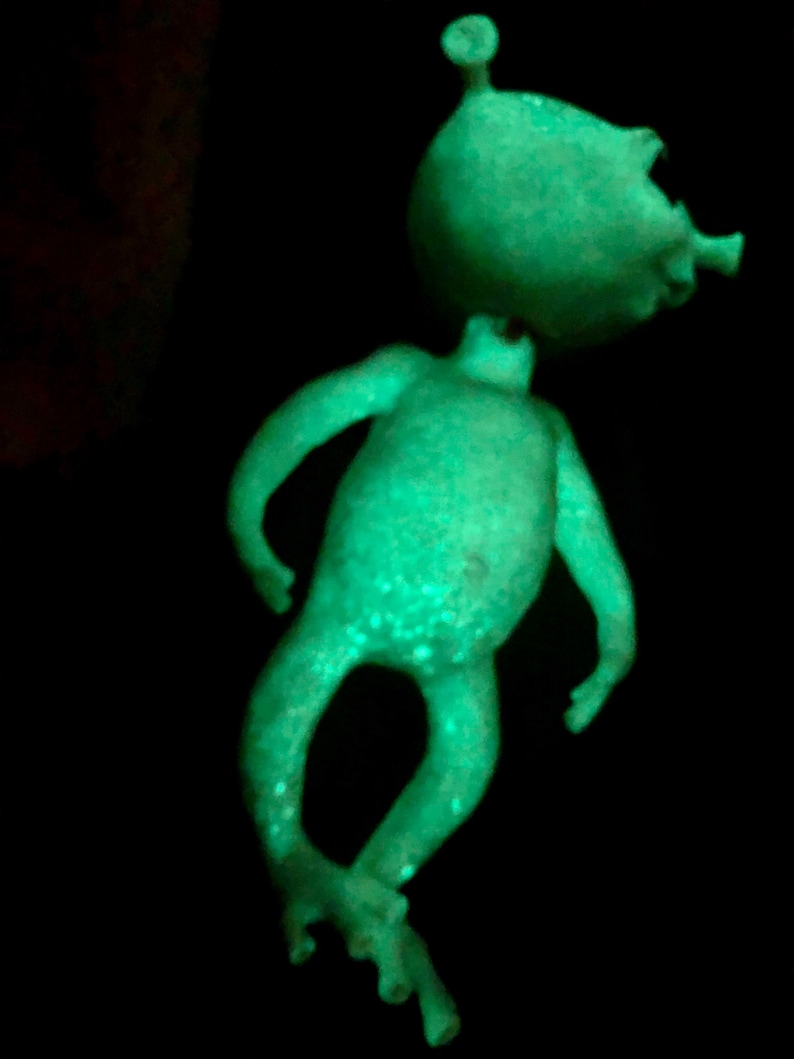 May include: A glowing green alien figurine with a large head and small body. The alien has a small antenna and is posed with its arms outstretched and legs crossed. The figurine has white speckles and is set against a black background.