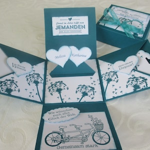 Könnte beinhalten: Tealblaue Explosionsbox mit weißem Hintergrund. Die Box ist mit Distel-Designs und dem Text "Einmal im Leben trifft man Jemanden der alles verändert." verziert. Die Box ist geöffnet und zeigt zwei Herzen mit den Namen "Melissa" und "Katharina". Die Box hat auch ein kleines Etikett mit dem Text "dem neuen Ehepaar!"
