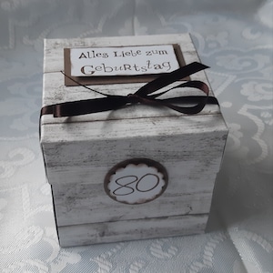 Könnte beinhalten: Eine weiße Geschenkbox mit Holzmaserung. Die Box ist mit einem braunen Band gebunden und hat eine braune Karte mit dem Text "Alles Liebe zum Geburtstag" und einen braunen Kreis mit der Nummer "80" darauf.