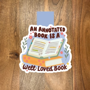 May include: A sticker featuring an open book with annotations, resting on a stack of books. The sticker has the text "AN ANNOTATED BOOK IS A Well-Loved Book" in brown and white, with floral and leaf accents.