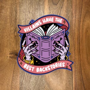 May include: A sticker featuring a purple book held by skeletal hands with long, pink nails. The book is open, with white pages. A banner above reads "Villains Have The", and below, "Best Backstories". The sticker has a black background with stars.