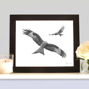 May include: Black and white framed art print featuring two birds of prey in flight. The larger bird is in the foreground with wings spread wide. The artwork is signed in the lower right corner. A candle and a flower are in the foreground.