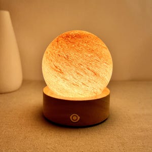 Może przedstawiać: Świecąca, kulista lampa z pomarańczowo-białym, marmurkowym szklanym kloszem stoi na okrągłej, drewnianej podstawie. Podstawa ma jeden, okrągły przycisk. Biały wazon w kształcie łzy znajduje się w tle.