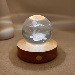 Op de afbeelding: Een heldere glazen bol met een geëtst ontwerp van een schildpad die zwemt onder een maan en wolken. De bol staat op een ronde, lichtgekleurde houten basis met een warme gloed. Een decoratief accent, ideaal voor een nachtkastje.