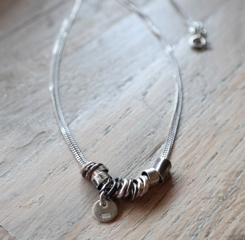 May include: A silver chain necklace with a silver charm featuring multiple intertwined rings and a small round tag with the inscription ".925".